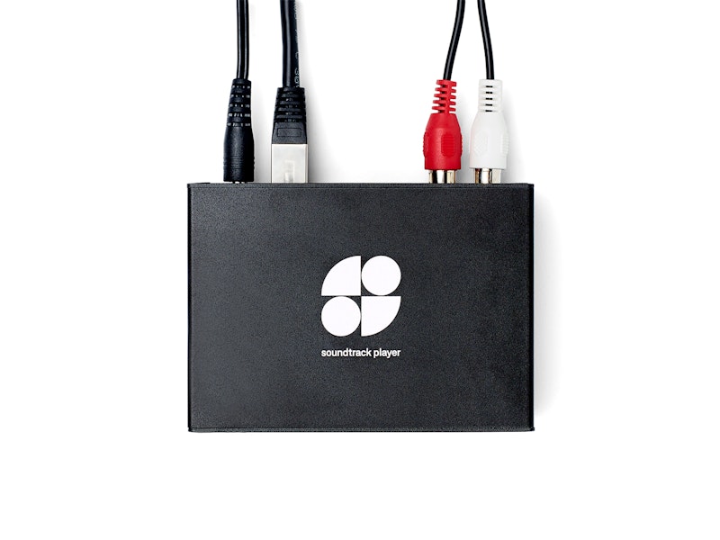 A black device labeled "soundtrack player" with a white logo consisting of four shapes arranged in a square pattern. The device has multiple cables connected to it, including a USB cable, an HDMI cable, and two RCA audio cables (one red and one white).