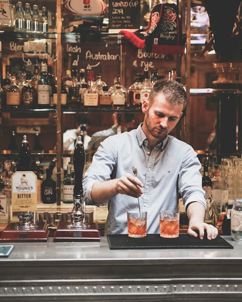 A bartender is preparing two cocktails behind a well-stocked bar, featuring a variety of liquor bottles.