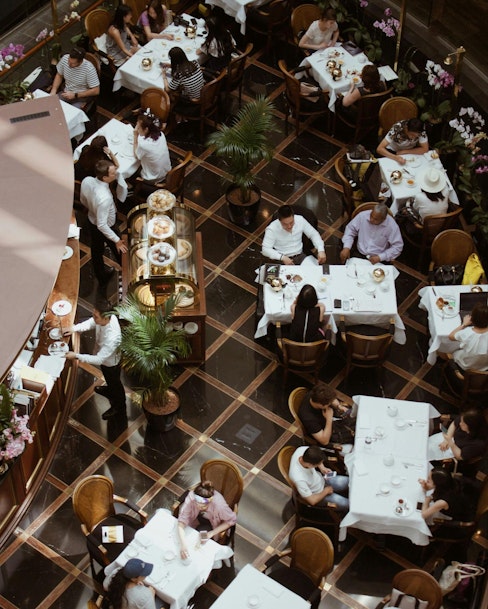 An aerial view of a busy restaurant with patrons seated at tables, enjoying their meals. The scene features waitstaff serving food and a central display of pastries.