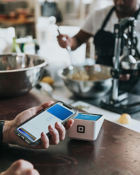 A person is holding a smartphone with a digital wallet app open, ready to make a payment using a Square contactless card reader. In the background, a chef is mixing ingredients in a large bowl.